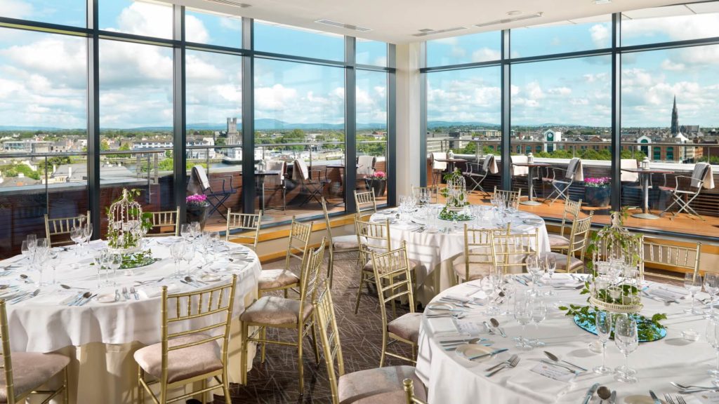 Elegant dining room with round tables set for an event, gold chairs, large windows, and a cityscape view with a terrace outside.