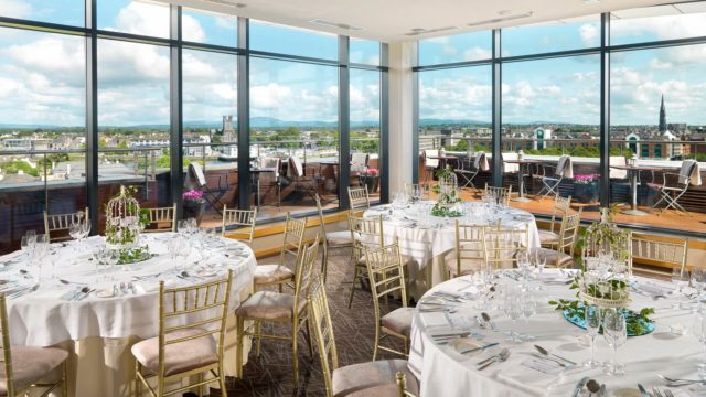 Elegant dining room with round tables set for an event, gold chairs, large windows, and a cityscape view with a terrace outside.
