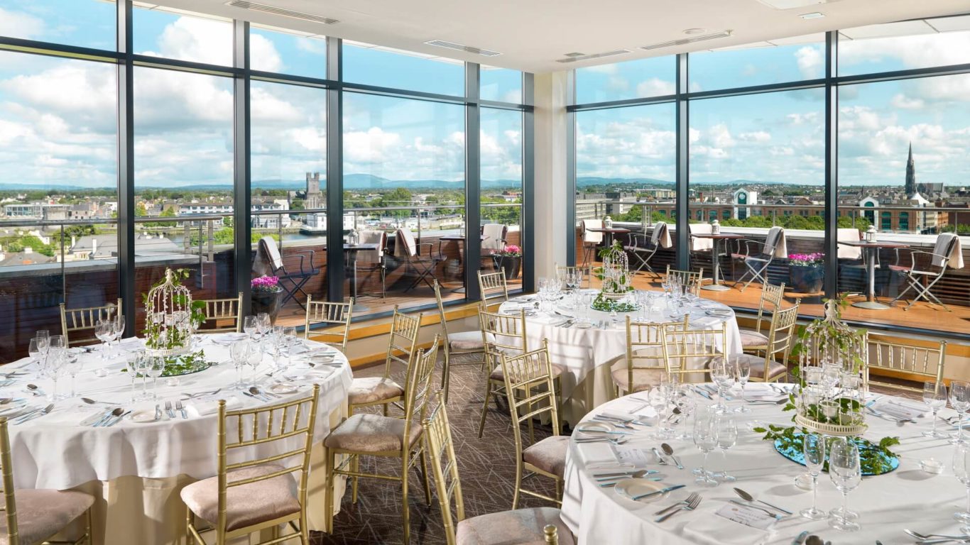 Banquet room with round tables set for an event, gold chairs, large windows, and a balcony overlooking a cityscape and distant hills under a partly cloudy sky.