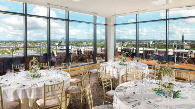 Bright banquet room with round tables set for an event, gold chairs, large windows, and a view of city buildings and a cloudy sky outside.