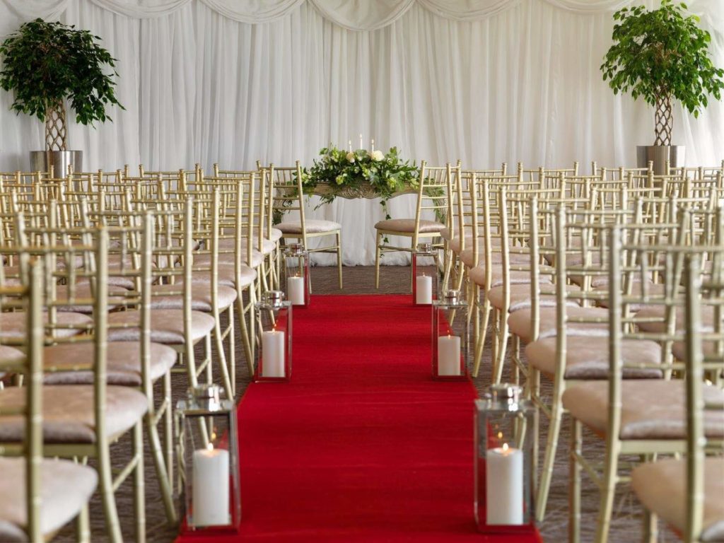 Rows of gold chairs face a table with floral arrangements at the end of a red carpet aisle, flanked by floor lanterns, in a white-draped indoor wedding venue.