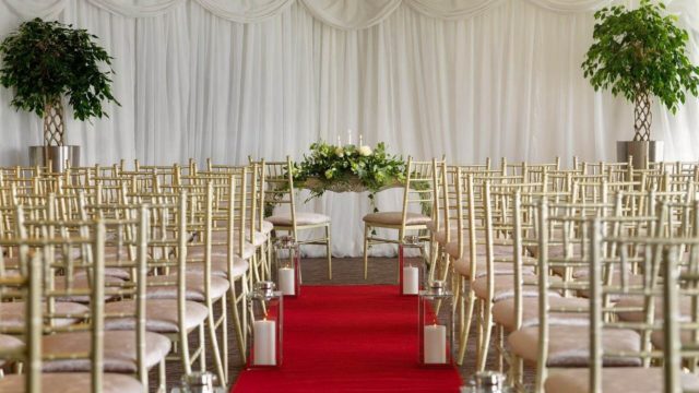 Rows of gold chairs facing a floral arrangement at the front, with a red carpet aisle and white draped backdrop, set for a formal indoor event or ceremony.
