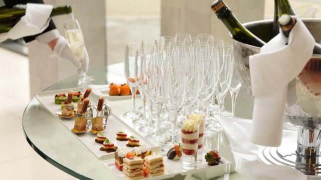 A table set with empty champagne glasses, assorted hors d'oeuvres on trays, bottles of champagne in an ice bucket, and a person pouring a drink.
