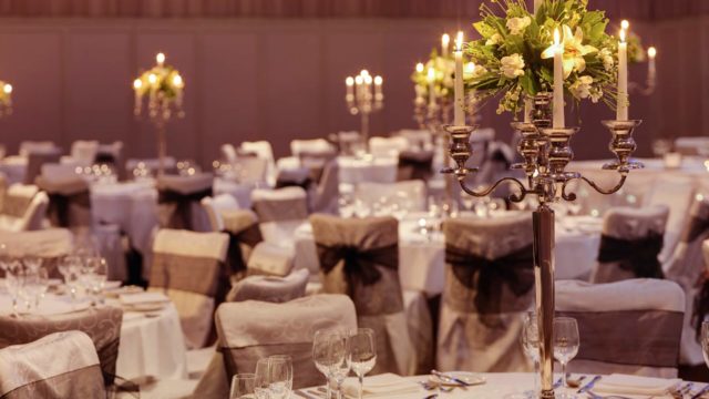 Round banquet tables with white linens, elegant place settings, and tall candelabra centerpieces decorated with flowers, set up for a formal event in a spacious hall.