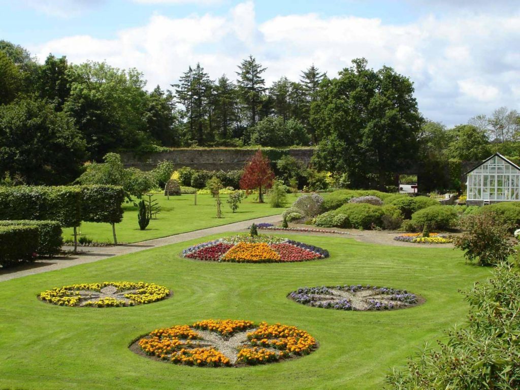 A landscaped garden with circular flower beds, neatly trimmed grass, hedges, a stone wall, and a greenhouse in the background under a partly cloudy sky.