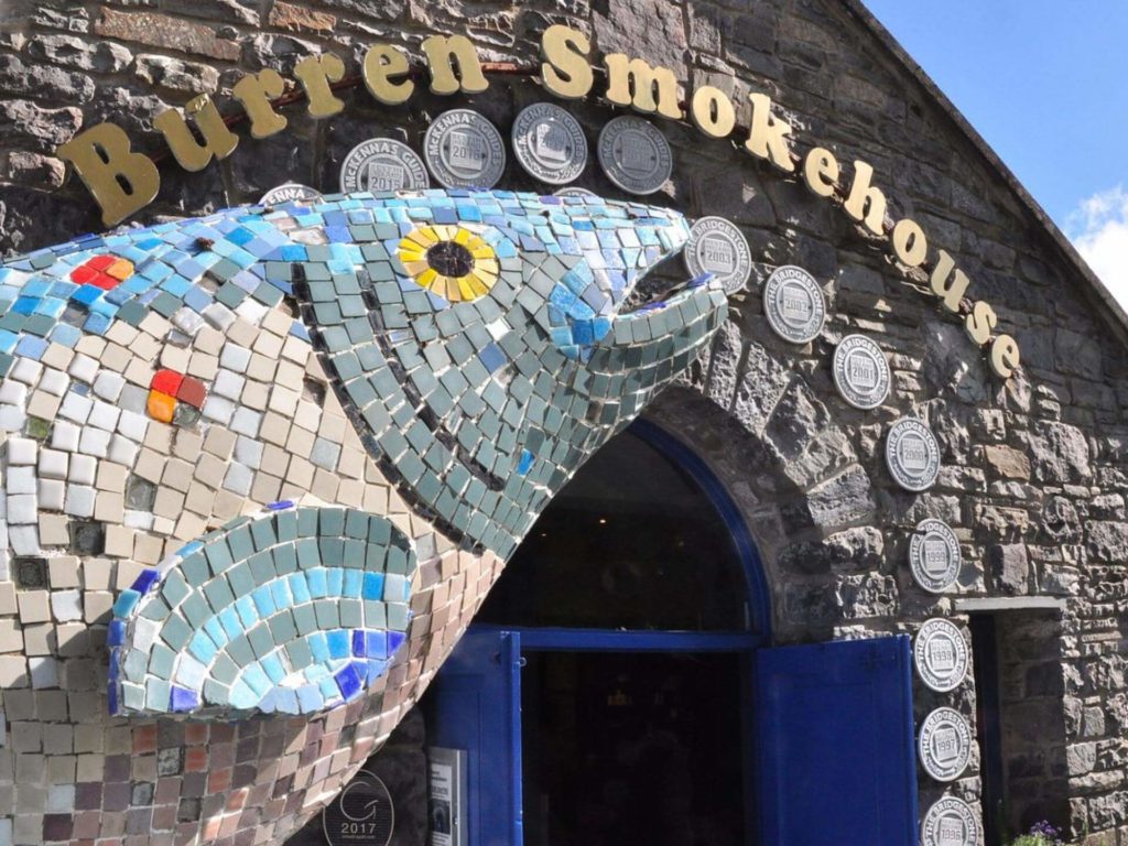 A mosaic fish sculpture decorates the entrance of Burren Smokehouse, a stone building with blue doors and award plaques displayed on the facade.