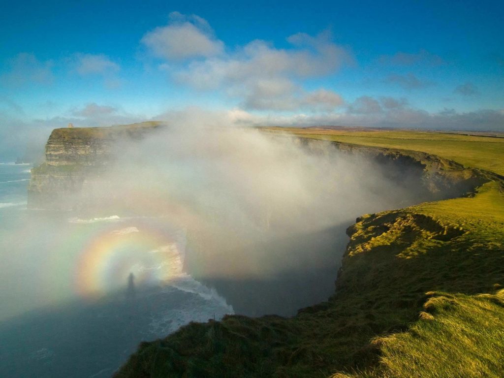 A coastal cliff covered in green grass with mist rising from the ocean below; a circular rainbow and the shadow of a person are visible in the fog.