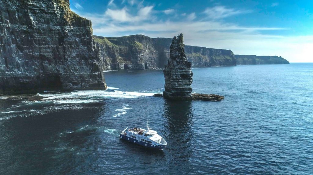 A tour boat sails near a tall sea stack and steep cliffs along a rocky coastline under a blue sky.
