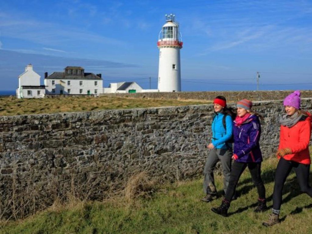 Three people in colorful outdoor clothing walk along a stone wall, with a lighthouse and white buildings in the background under a blue sky.