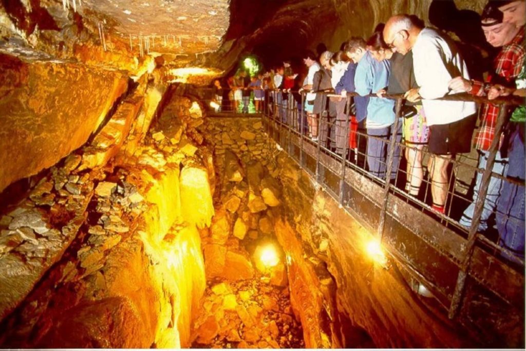 A group of people stands on a walkway inside an illuminated cave, observing the rocky formations and crevice below.