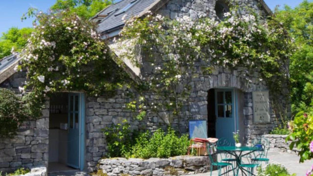 A stone cottage with flowering vines on the walls, blue doors, and a small outdoor table and chairs set on a sunny day.