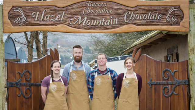 Four people wearing brown aprons stand under a wooden sign that reads 