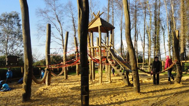 Children play on a wooden playground structure with ropes and slides in a sandy outdoor area surrounded by tall trees.