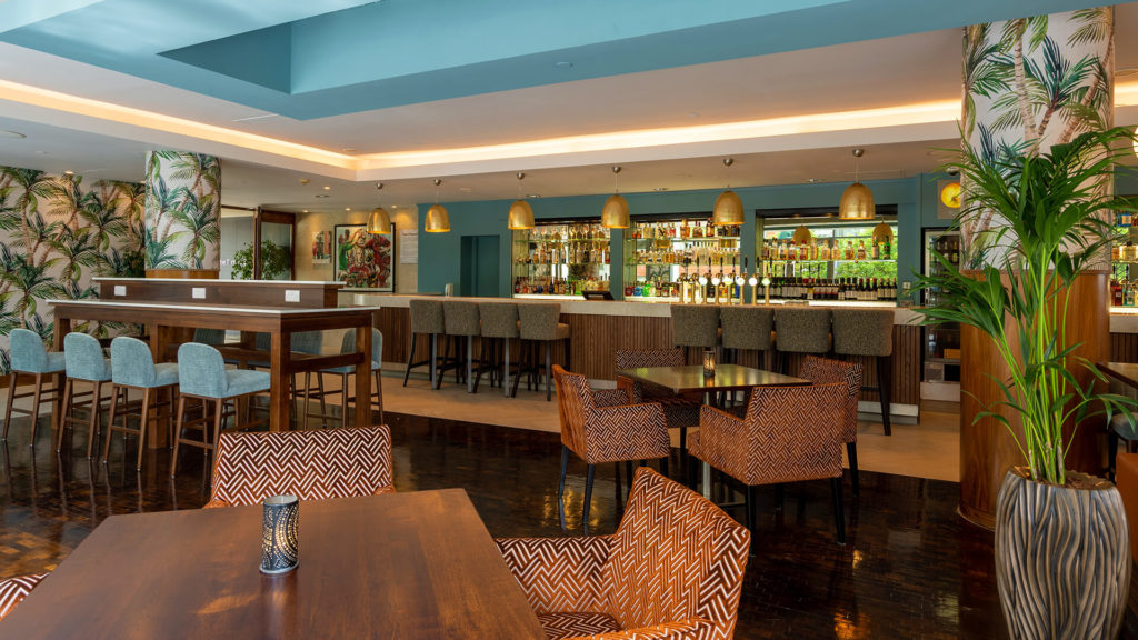 A modern bar and lounge area featuring a long bar with high stools, multiple tables and chairs, tropical plants, and decorative lighting.