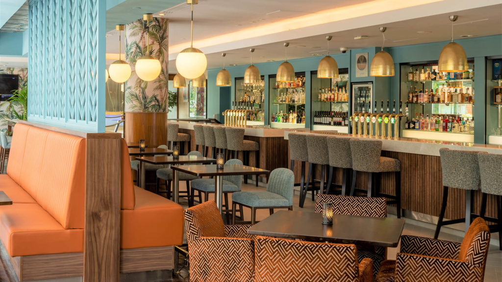 Modern bar with upholstered seating, patterned chairs, wooden tables, and a long counter lined with barstools; shelves stocked with bottles and hanging globe pendant lights.