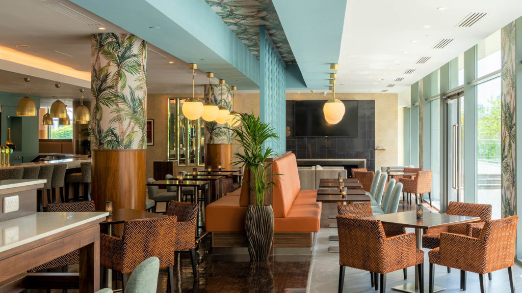 Modern restaurant interior with patterned chairs, a long orange booth, large windows for natural light, and decorative plants. Warm lighting and tropical-themed decor elements are present.
