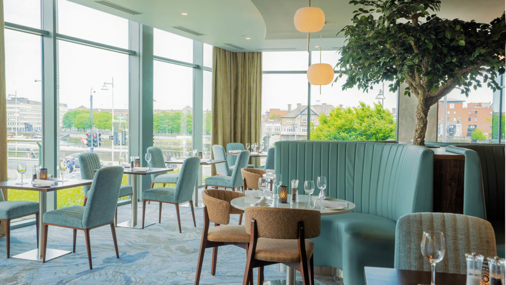 Modern restaurant interior with large windows, light blue booth seating, wooden chairs, set tables with glassware, and a decorative indoor tree, overlooking a cityscape.