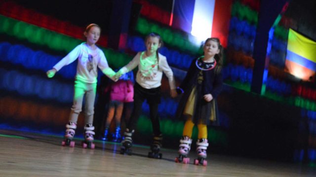Three young children roller skate indoors, holding hands, with blurred colorful lights and flags in the background.