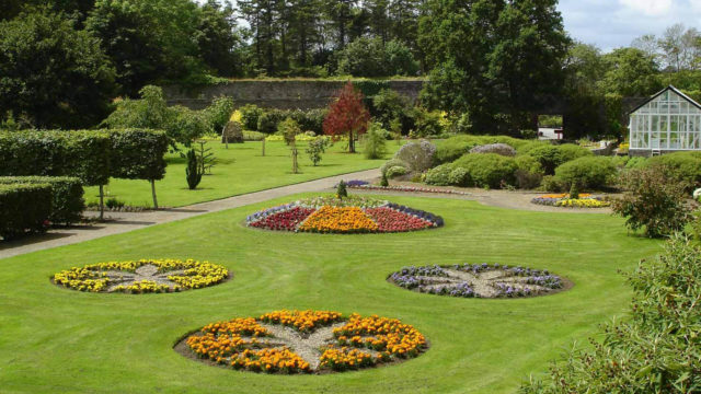A formal garden features circular flower beds with colorful blooms, neatly trimmed hedges, a greenhouse, and a background of trees and stone walls.