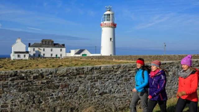 Three people in colorful outdoor clothing walk along a stone wall with a lighthouse and adjacent white buildings in the background under a blue sky.