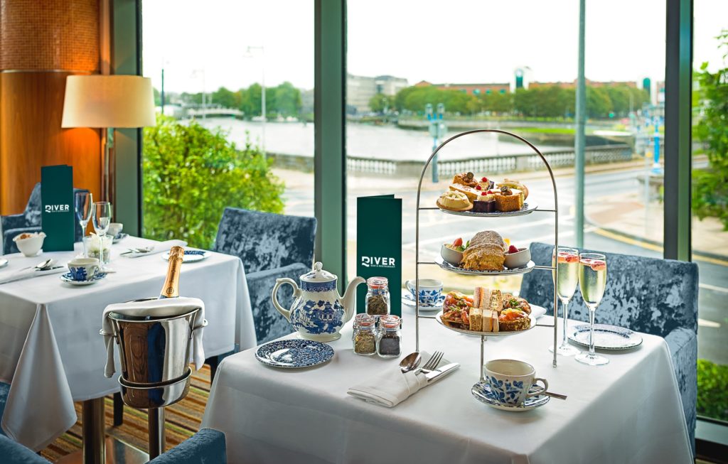 A table set for afternoon tea with a tiered tray of pastries and sandwiches, fine china, two glasses of champagne, and a river view through large windows in the background.