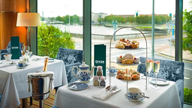 A table set for afternoon tea with a tiered tray of pastries and sandwiches, fine china, two glasses of champagne, and a river view through large windows in the background.