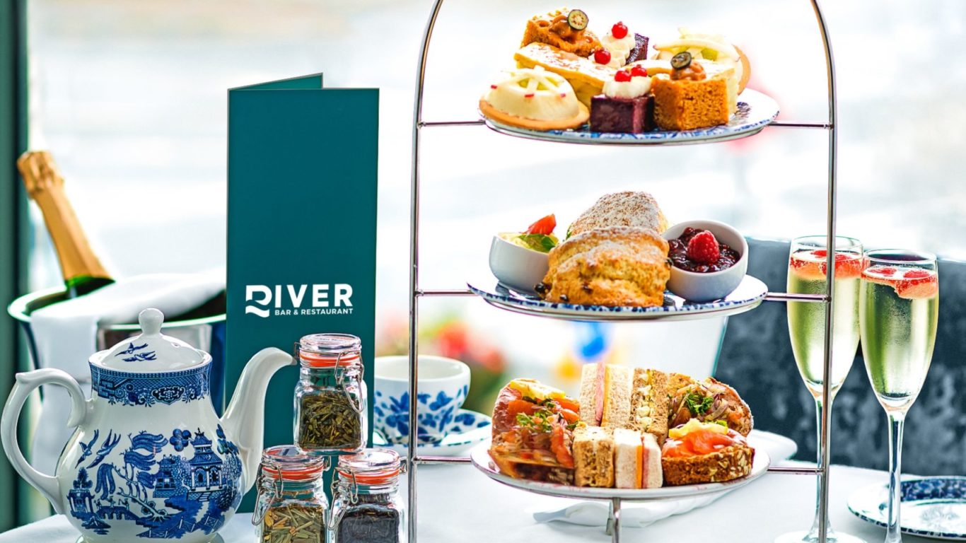 A table set for afternoon tea with a teapot, teacups, tiered stand of pastries and sandwiches, two glasses of champagne, and a menu, by a window with a river view.