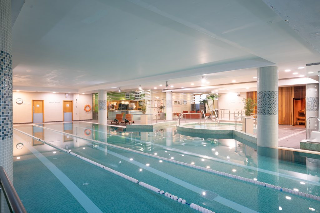 Indoor swimming pool with marked lanes, a round Jacuzzi, lounge chairs, and sauna room doors in a well-lit spa facility.