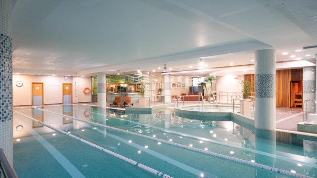 Indoor swimming pool with marked lanes, a round Jacuzzi, lounge chairs, and sauna room doors in a well-lit spa facility.