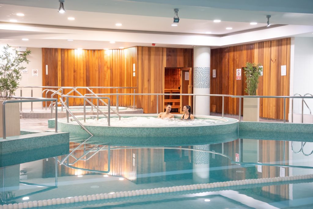 Two people are relaxing in a circular hot tub next to an indoor pool, with wooden paneling and a sauna visible in the background—perfect for cozy Autumn Hotel Breaks.