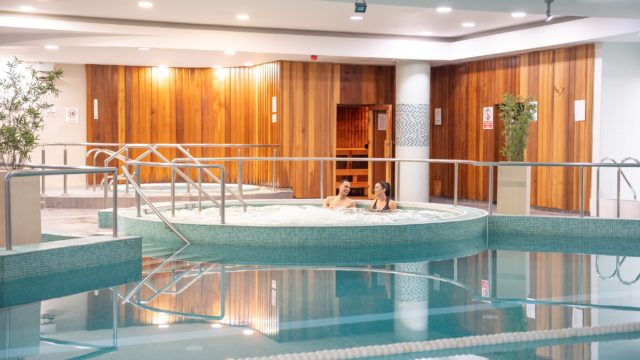 Two people are relaxing in a circular hot tub next to an indoor pool, with wooden paneling and a sauna visible in the background—perfect for cozy Autumn Hotel Breaks.
