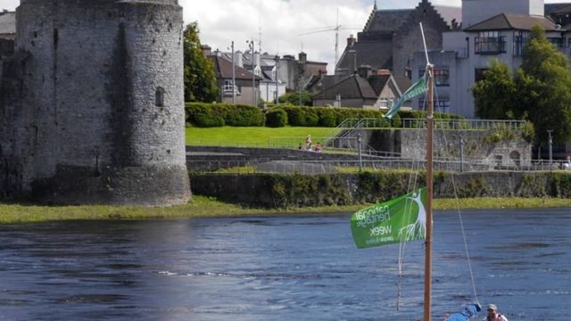 A group of people sails a small boat with green flags on a river; a stone castle with a red flag and buildings are visible in the background.