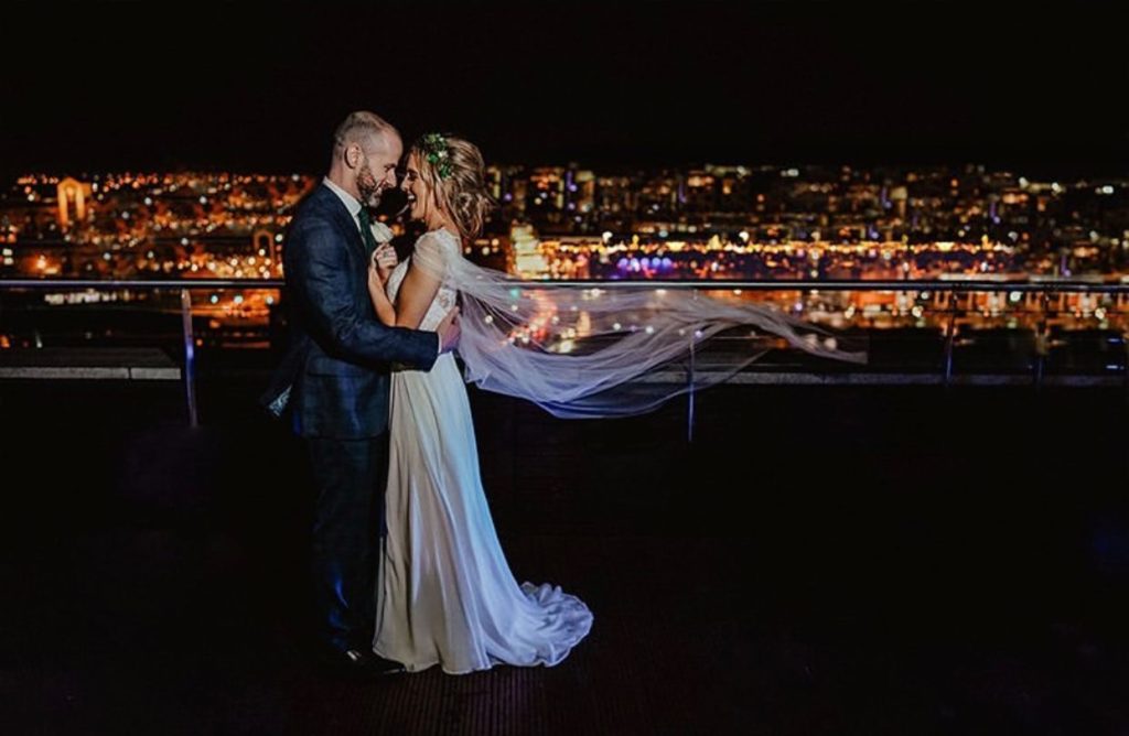 A bride and groom stand close together on a rooftop at night, with city lights glowing in the background and the bride's veil blowing in the wind.