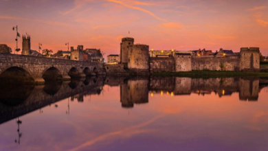 A stone bridge and medieval castle are reflected in a calm river at sunset, with purple and orange clouds in the sky&mdash;an enchanting scene perfect for those seeking idyllic Autumn Hotel Breaks.