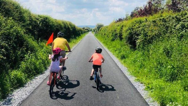 Three people wearing helmets ride bicycles on a paved path bordered by green bushes under a partly cloudy sky.