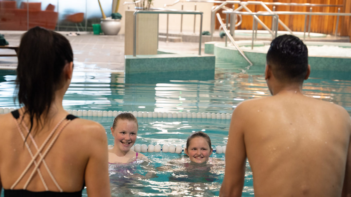 Two adults and two children enjoy an indoor swimming pool during their Autumn Hotel Break, with the smiling children looking at the adults across a lane divider.
