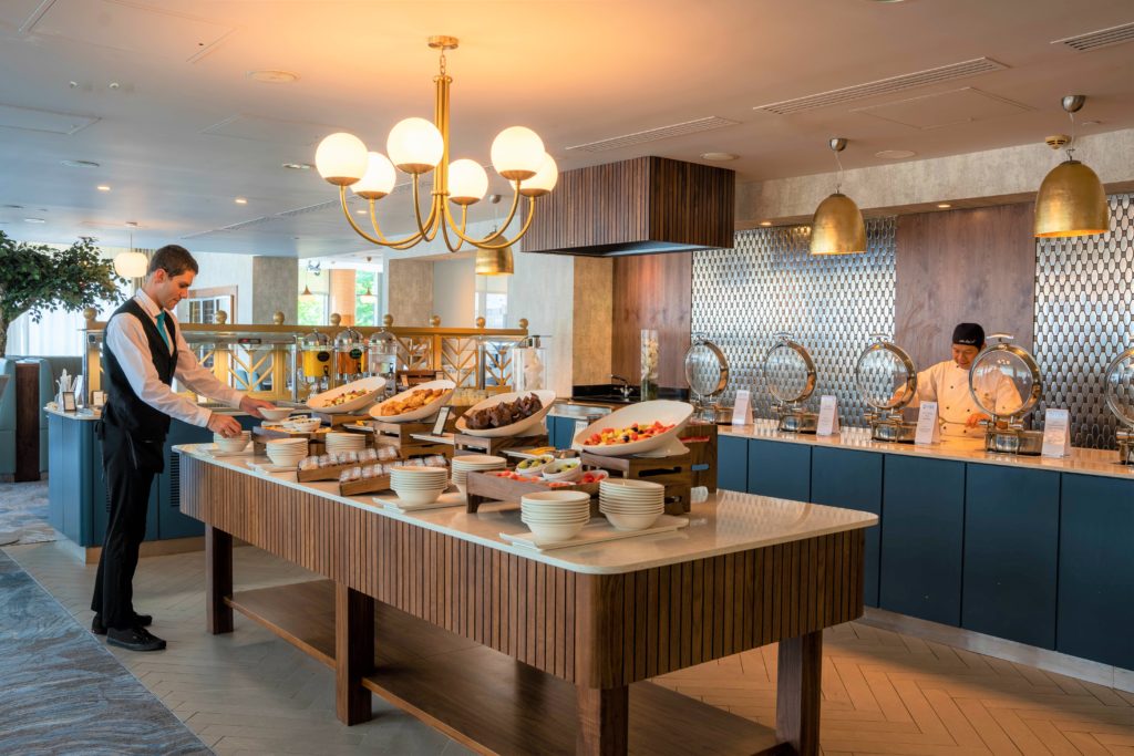 A buffet setup in a restaurant with a waiter arranging food on a central table and a chef standing behind warming trays on the counter.