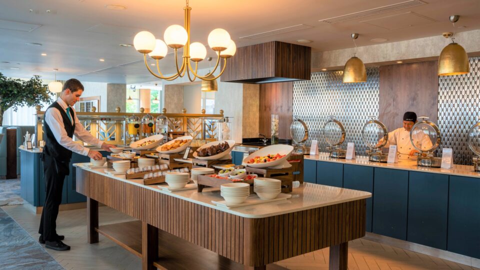 A buffet setup in a restaurant with a waiter arranging food on a central table and a chef standing behind warming trays on the counter.