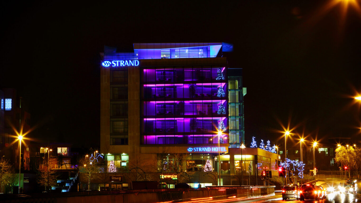 A multi-story hotel with purple-lit balconies and STRAND HOTEL signage is illuminated at night, with streetlights and car light trails visible in the foreground.