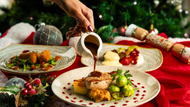 A hand pours gravy onto a plate with roast meat, Brussels sprouts, potatoes, and stuffing, set on a festive table with Christmas decorations in the background.