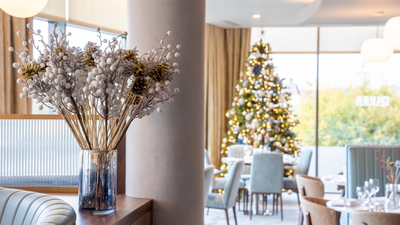 A modern restaurant interior with a glass vase of decorative white branches in the foreground and a decorated Christmas tree in the background.