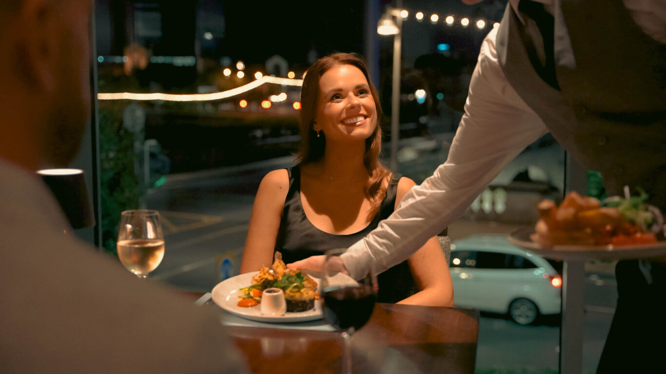 A woman sits at a restaurant table with food and wine, smiling at a server who is placing a plate in front of another diner. Evening city lights twinkle through the window, setting the perfect mood for Valentine's in Limerick.