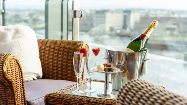 A wicker chair beside a glass table with two champagne flutes, chocolate truffles, and a bottle of champagne in an ice bucket—perfect for celebrating Valentine's in Limerick while overlooking the cityscape.