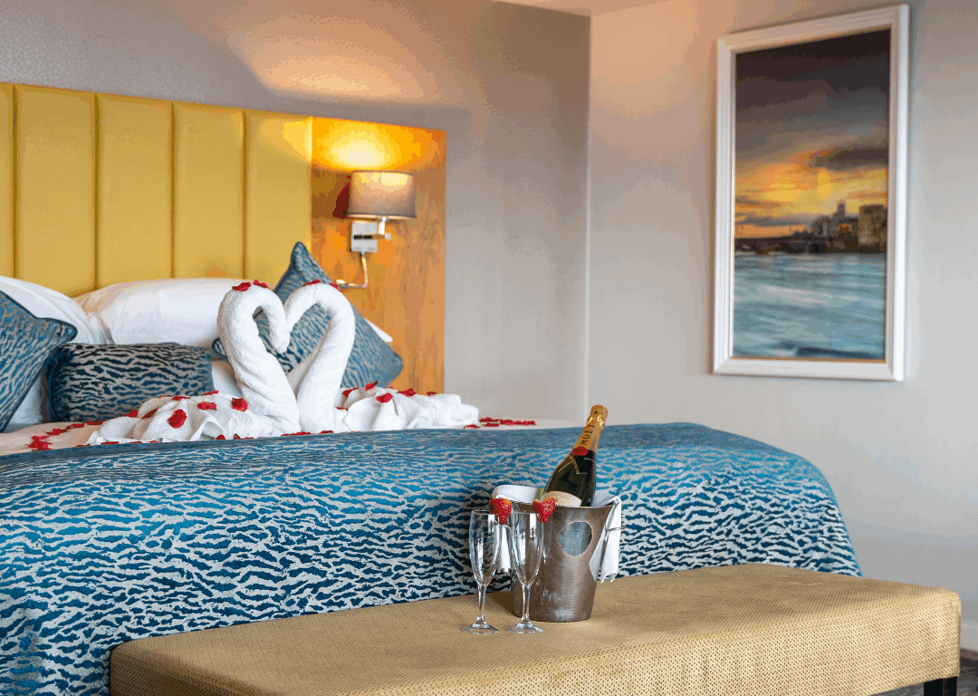 A hotel room features a neatly made bed with towel swans, two champagne glasses and a bottle in an ice bucket, a cushioned bench, and a framed picture on the wall.