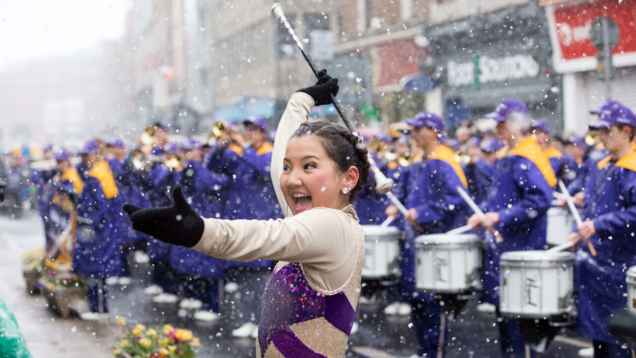 A baton twirler in a sparkly outfit performs in front of a marching band dressed in purple uniforms during a snowy outdoor parade.