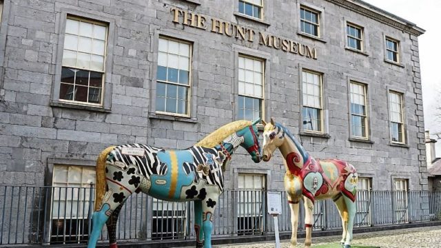 Two colorful, patterned horse statues stand in front of the stone facade of The Hunt Museum, which has several windows and a metal fence.