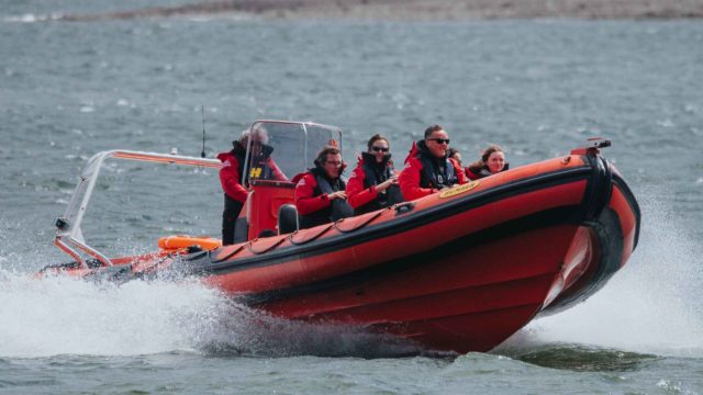 Five people in red jackets ride a fast-moving orange inflatable boat on a body of water, with spray rising from the boat's sides.