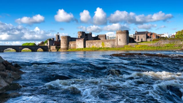 A stone castle with round towers stands beside a river, with a stone bridge to the left and blue sky with clouds above.