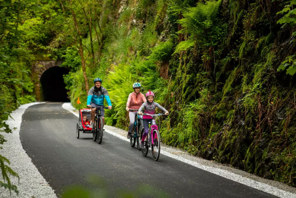 Three people ride bicycles on a paved path through a lush, green forest during a Summer Family Break; one pulls a child trailer. A tunnel entrance is visible in the background.