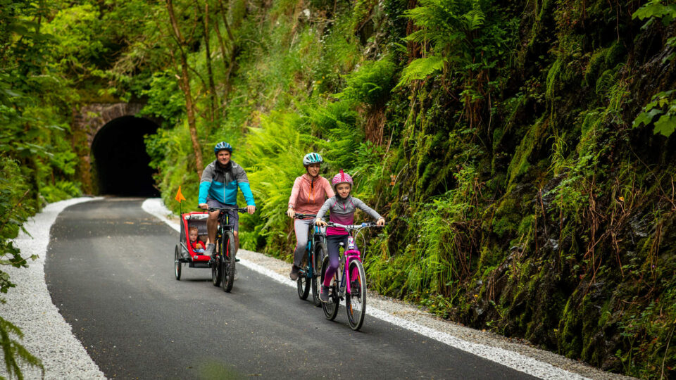 Three people ride bicycles on a paved path through a lush, green forest during a Summer Family Break; one pulls a child trailer. A tunnel entrance is visible in the background.